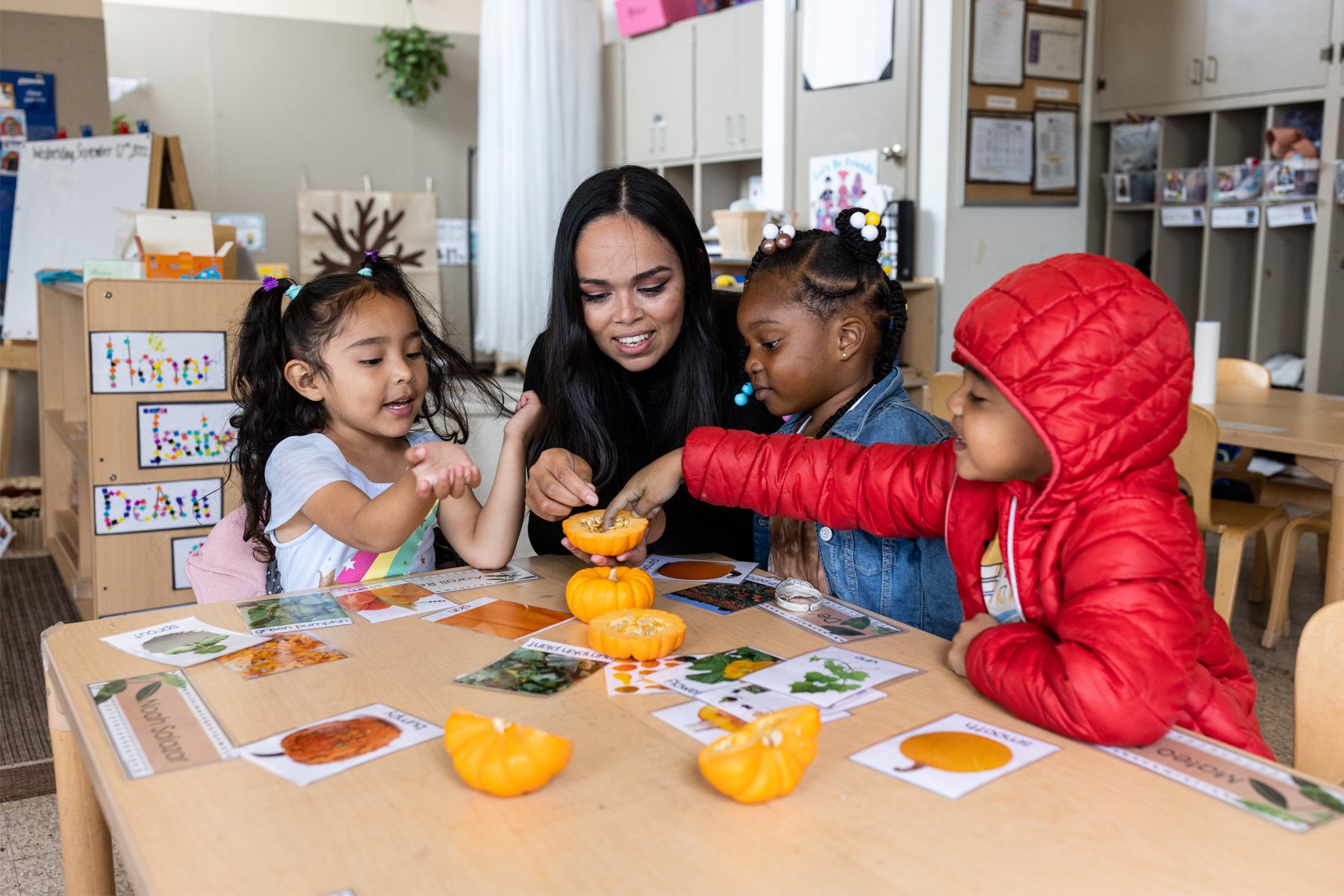 Three young children play with a miniature pumpkin at a table with a teacher.