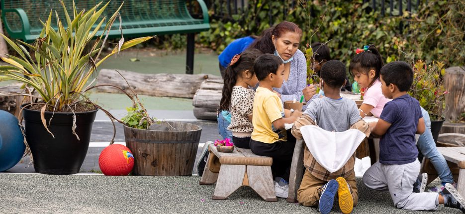 Young children gather at a table outside with teacher.