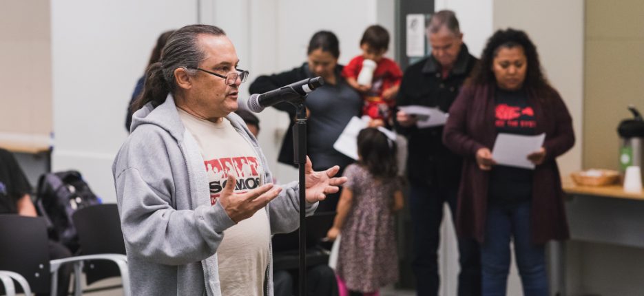 A man standing at a microphone speaking, while others wait in line.