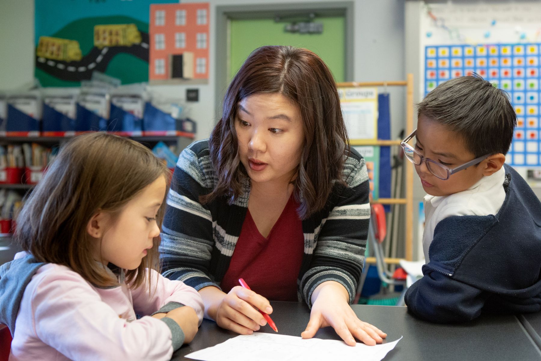 A woman speaking to two young children at a table, while writing on a piece of paper.