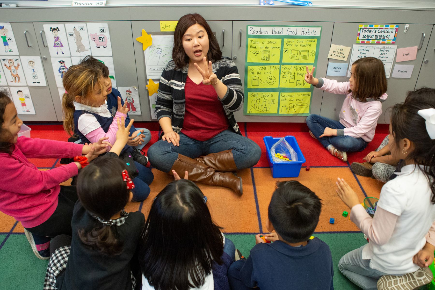 A woman sits on the ground in a circle with a group of young children counting on fingers.