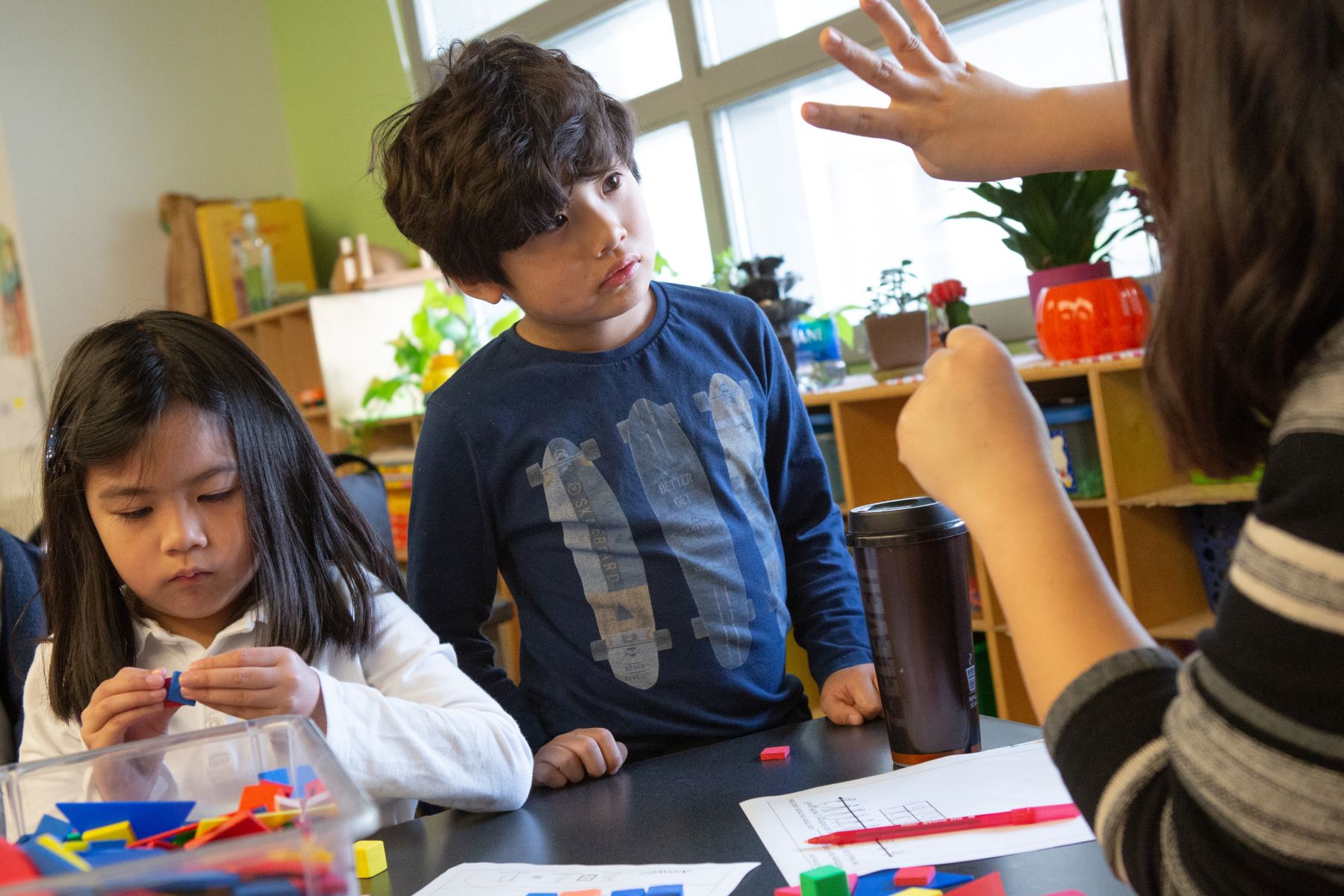 A young boy looking at an adult counting with fingers, while a young girl plays with blocks.