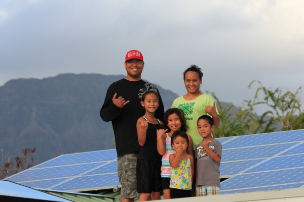 Two adults and four kids smile at camera in front of solar panels.